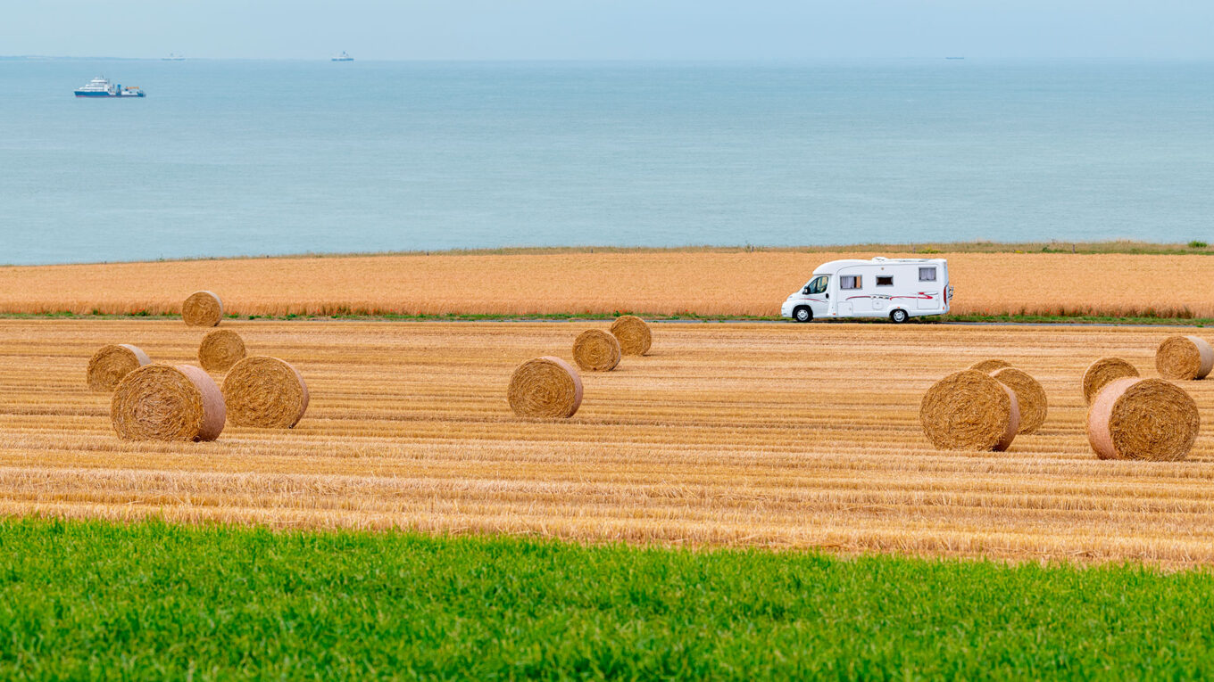 Autocamper kører forbi halmballer på mark med udsigt til havet i Normandiet