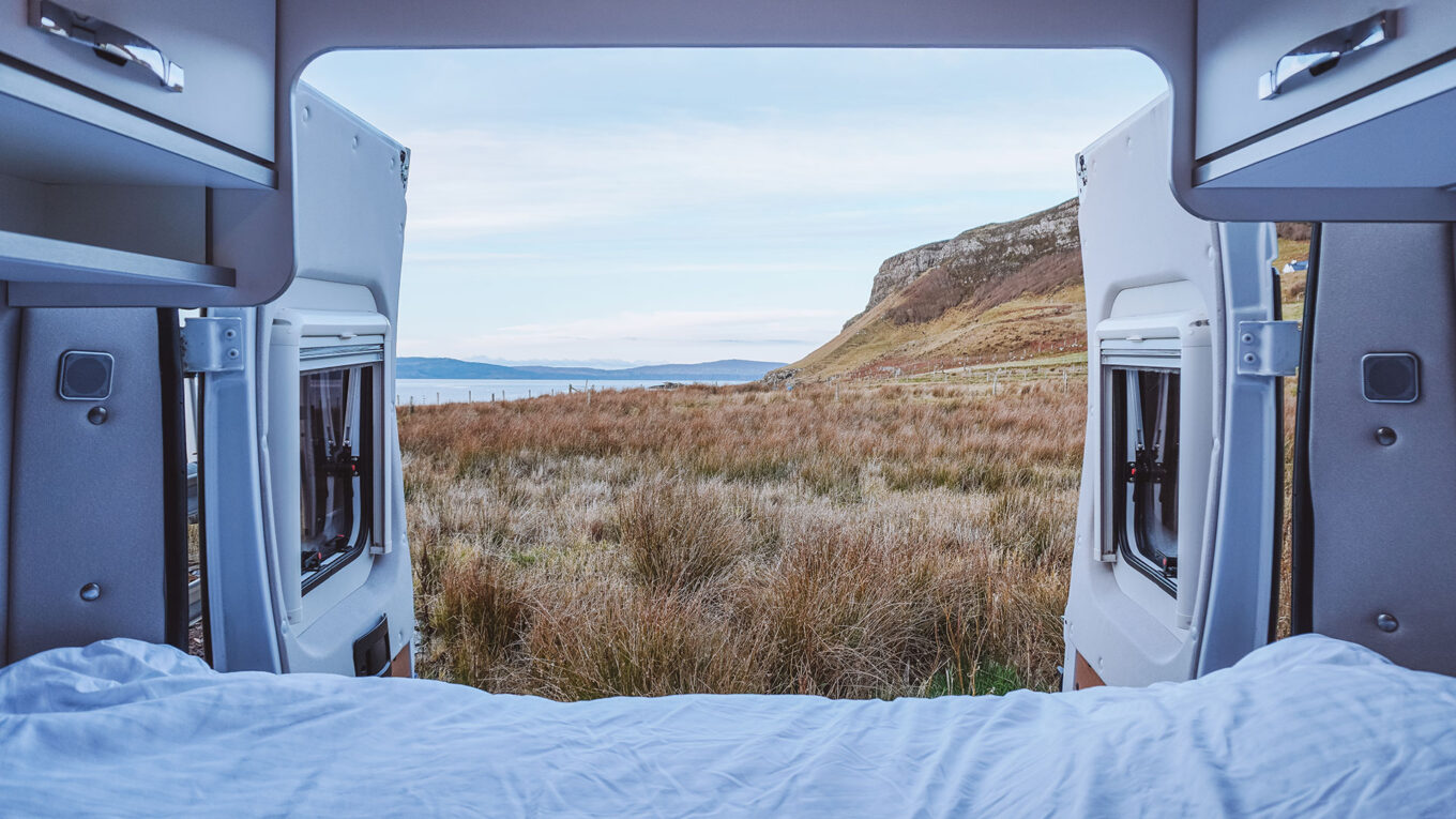 Indvendigt kig ud fra en campervan med åbne bagdøre og panoramaudsigt til skotsk natur – afslappende moment på autocamperferie.